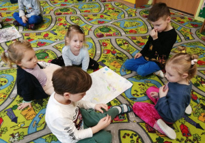 Dzieci siedzą na dywanie i pokazują pracę plastyczną